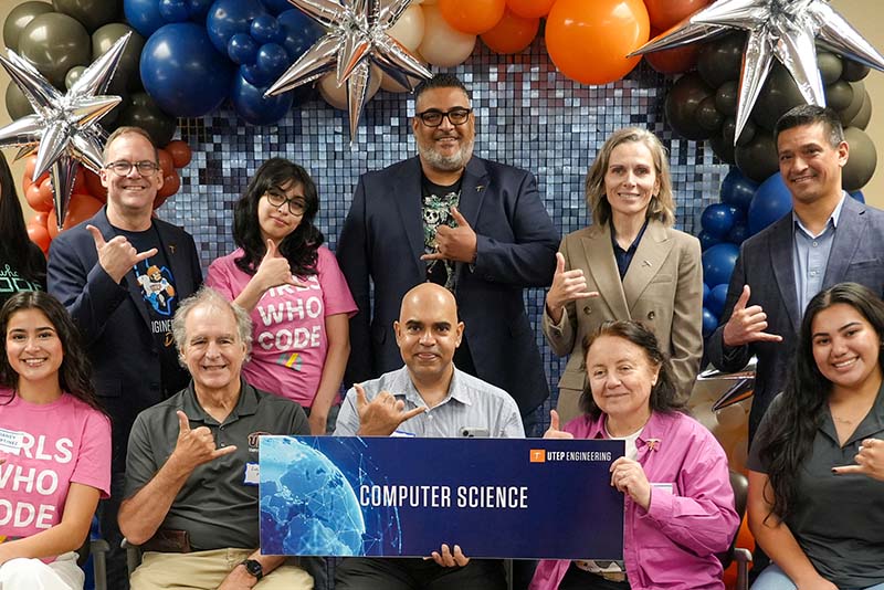 Group of attendees pose together during a UTEP Engineering homecoming gathering while holding a large “Industrial, Manufacturing and Systems Engineering” banner, with participants seated and standing in front of a backdrop decorated with orange, blue, black, and silver balloons while making hand gesture signs for the group photo.